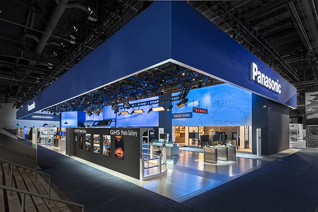 An angled view of a large trade show exhibit with a blue banner abover with a white "Panasonic" illunimated sign on each side and a blue and white interior with multiple exhibits and displays.