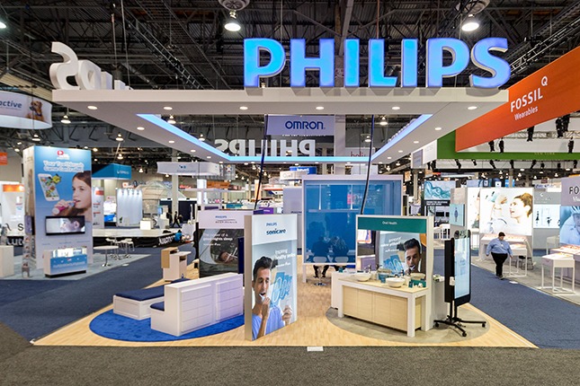 A trade show exhibit with an illuminated blue Philips logo suspended above upon a white soffit and multiple signs and displays beneath.