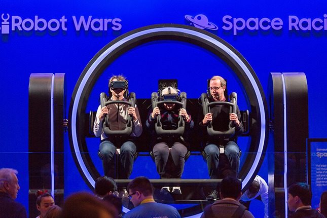 A three-person circular virtual reality ride with two men and an elderly woman seated in the center in front of a blue background which says Robot Wars and Space Racing and a small crowd in front.