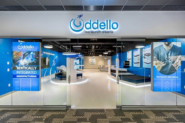 The exterior of the Oddello shop with glass walls and glass sliding doors and a white and blue interior with beds on display.
