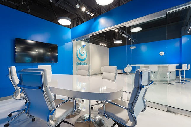 A conference room with blue walls and a white conference talbe with modern white chairs and a LCD TV on the wall and a white-and-blue retail area outside a glass door.