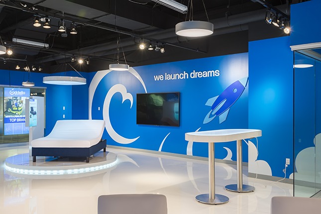 A blue wall in a retail space with graphic art of a blue rocket and white swirls with the text "we launch dreams" above and a bed on a pedestal and an illuminated table in the foreground.