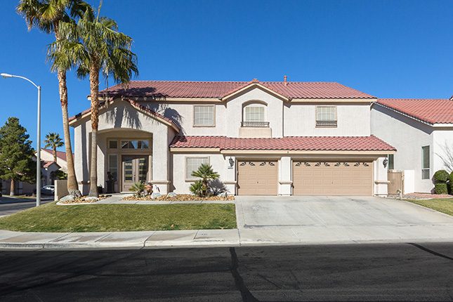 A white stucco home with a three-car garage, red-tile roof, gabled entry and two tall palm trees beneath a clear blue sky.