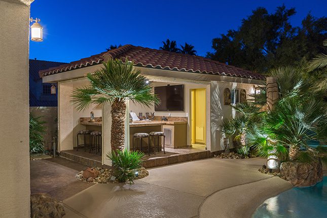 A white stucco pool house with an open kitchen and barstools visible and palm trees surrounding it with a deep blue sky and silhouettes of trees behind it.