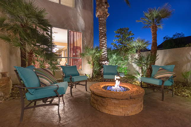 Four metal patio chairs with teal-colored cushions surrounding a fire pit at dusk with palm trees surrounding them and the living room of the home visible through a large window in the background.