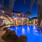 An electric b;ue illuminated swimming pool with rock landscaping and small waterslide visible through palm fronds in the foreground and a two-story home and pool house in the background.