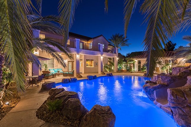 An electric b;ue illuminated swimming pool with rock landscaping and small waterslide visible through palm fronds in the foreground and a two-story home and pool house in the background.