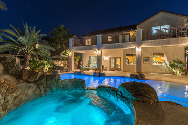 Azure water in rock-surrounded a hot tub pouring into an illuminated blue swimming pool at night with a two-story home in the background.