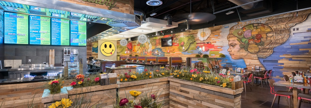 The entrance to a restaurant queue made of wood planters with faux grass and flowers atop which leads to a counter with LCD menus above. There are tables and booths off to the right in the background and a eclectic wall mural.