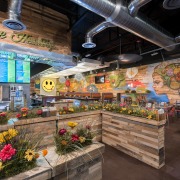 The entrance to a restaurant queue made of wood planters with faux grass and flowers atop which leads to a counter with LCD menus above. There are tables and booths off to the right in the background and a eclectic wall mural.