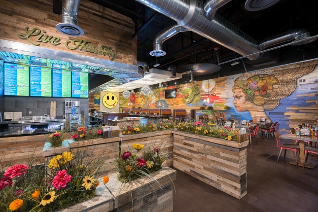 The entrance to a restaurant queue made of wood planters with faux grass and flowers atop which leads to a counter with LCD menus above. There are tables and booths off to the right in the background and a eclectic wall mural.