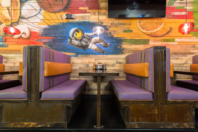 A small section of restaurant booths with purple cushions and a wall mural painted on wood of an astronaut with a smiley for a face on a blue background.