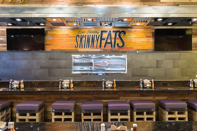 A straight-on shot of the skinnyfats dining counter with 8 purple-cushioned barstools in front and the window to the kitchen behind and a painted SkinnyFats logo above between two LCD TVs.