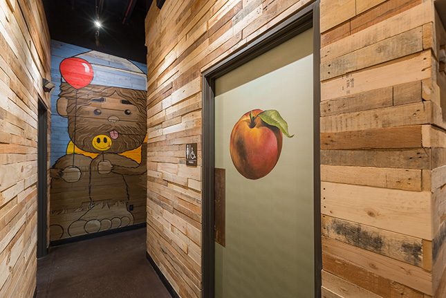 A hallways of exposed wood-plank walls with a ladies restroom door in the foreground with a peach painted on it and a blue-and-brown mural on the back wall of a bear liking a smiley-faced lollipop.