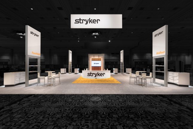 A centered photo of the Stryker booth at ACFAS 2017 with hanging banner, welcome counter with LCD screen, and four vertical signs all with the Stryker logo--edited so that background is dimmed.