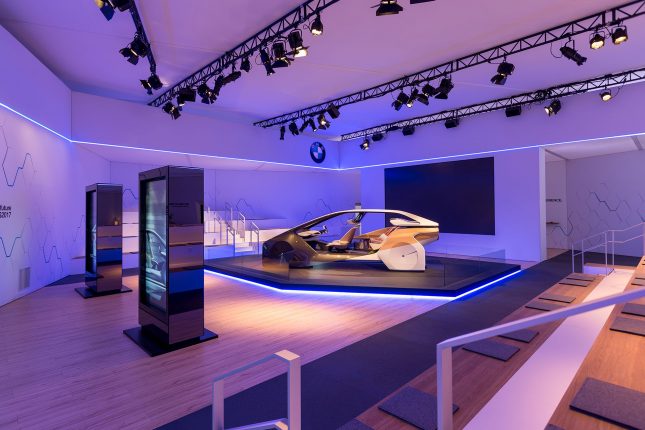 A white area with hardwood flooring and illuminated blue and purple with lighting, a BMW concept car on a pedestal and large black video screen in the background and two black vertical video kiosks in the foreground.
