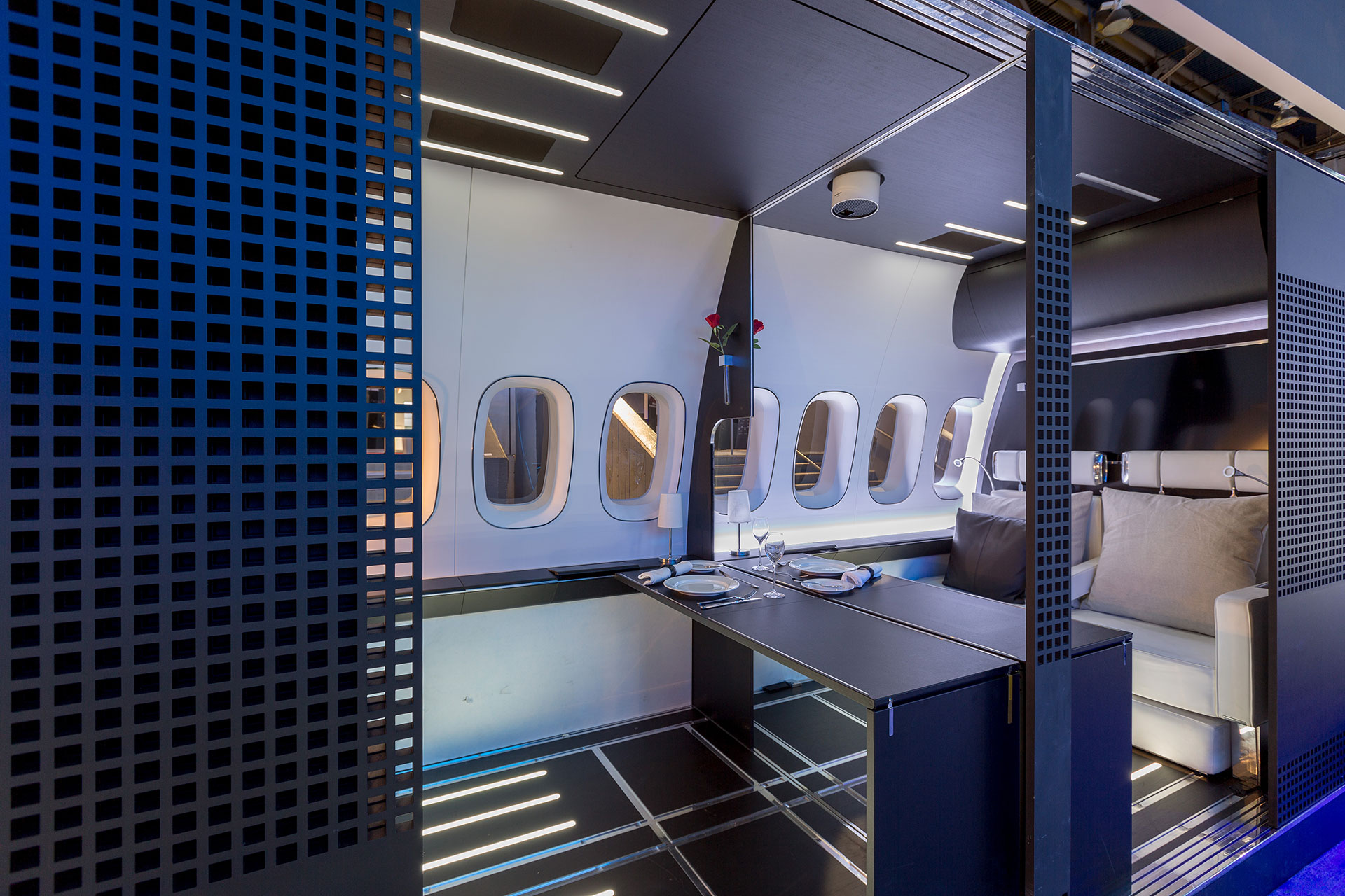 Replica of a First-class aircraft cabin as a trade show exhibit at CES 2016 with dinnerware laid out on a black table with white walls shot from the outside looking in.