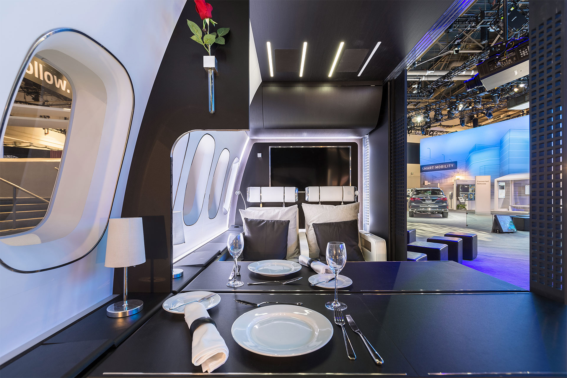 Replica of a First-class aircraft cabin as a CES 2016 trade show exhibit with dinnerware laid out on a black table with white walls shot from the perspective of a seated passenger.
