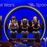 A three-person circular virtual reality ride with two men and an elderly woman seated in the center in front of a blue background which says Robot Wars and Space Racing and a small crowd in front.