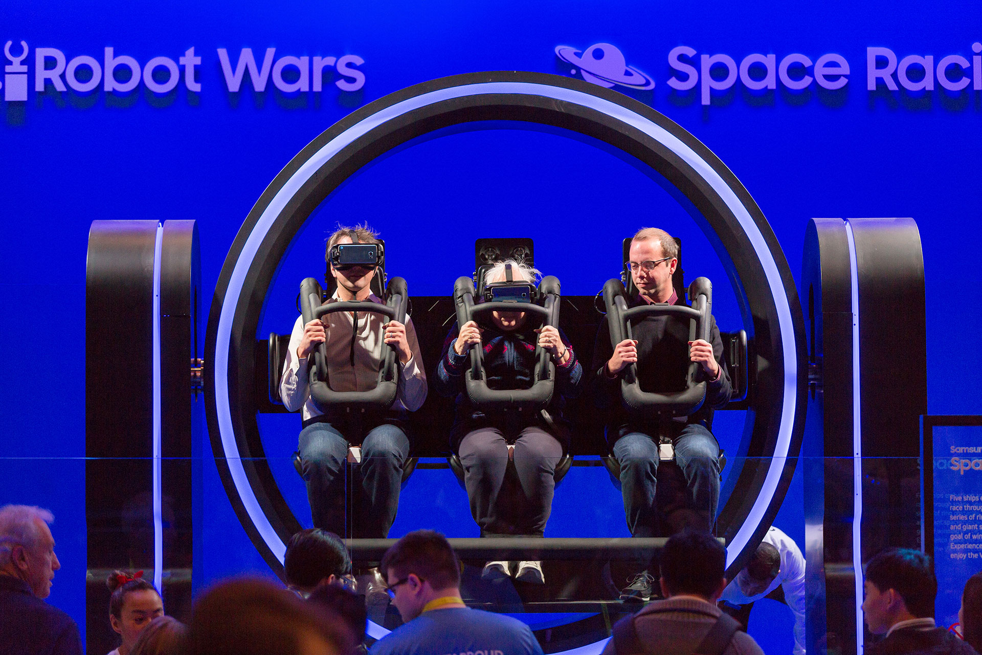 A three-person circular virtual reality ride with two men and an elderly woman seated in the center in front of a blue background which says Robot Wars and Space Racing and a small crowd in front.