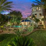 The backyard of a spanish-style villa with grass and desert foliage in the foreground, patio furniture and umbrella in the center and various trees including palm trees throughout and a pool and home in the background amidst a blue-and-red sunset.