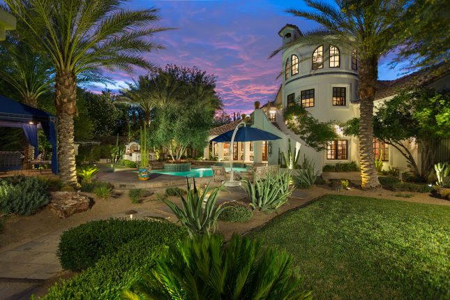 The backyard of a spanish-style villa with grass and desert foliage in the foreground, patio furniture and umbrella in the center and various trees including palm trees throughout and a pool and home in the background amidst a blue-and-red sunset.