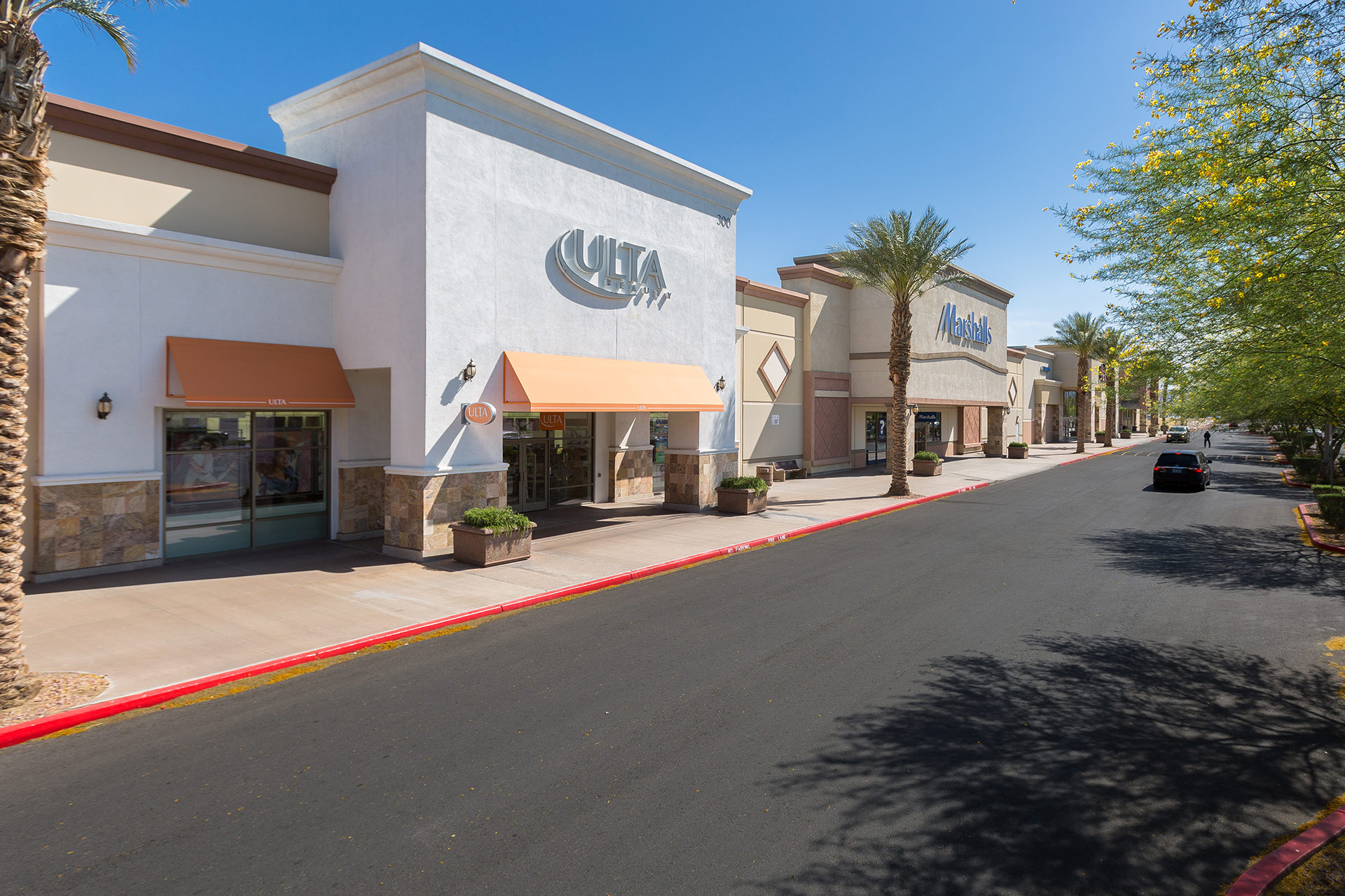 A shopping complex at midday with a white Ulta store in the foreground and beige Marshalls beside it and other retailers in the distance and cars driving in front of it.
