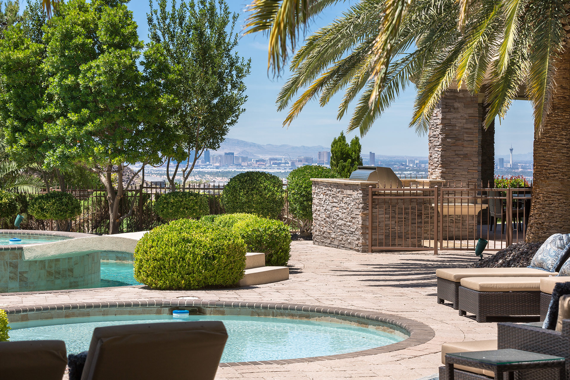 A wading pool, backyard grill with stone enclosure, palm fronds int he foreground, hedges and trees about and a daytime view of the Las Vegas strip in the background.