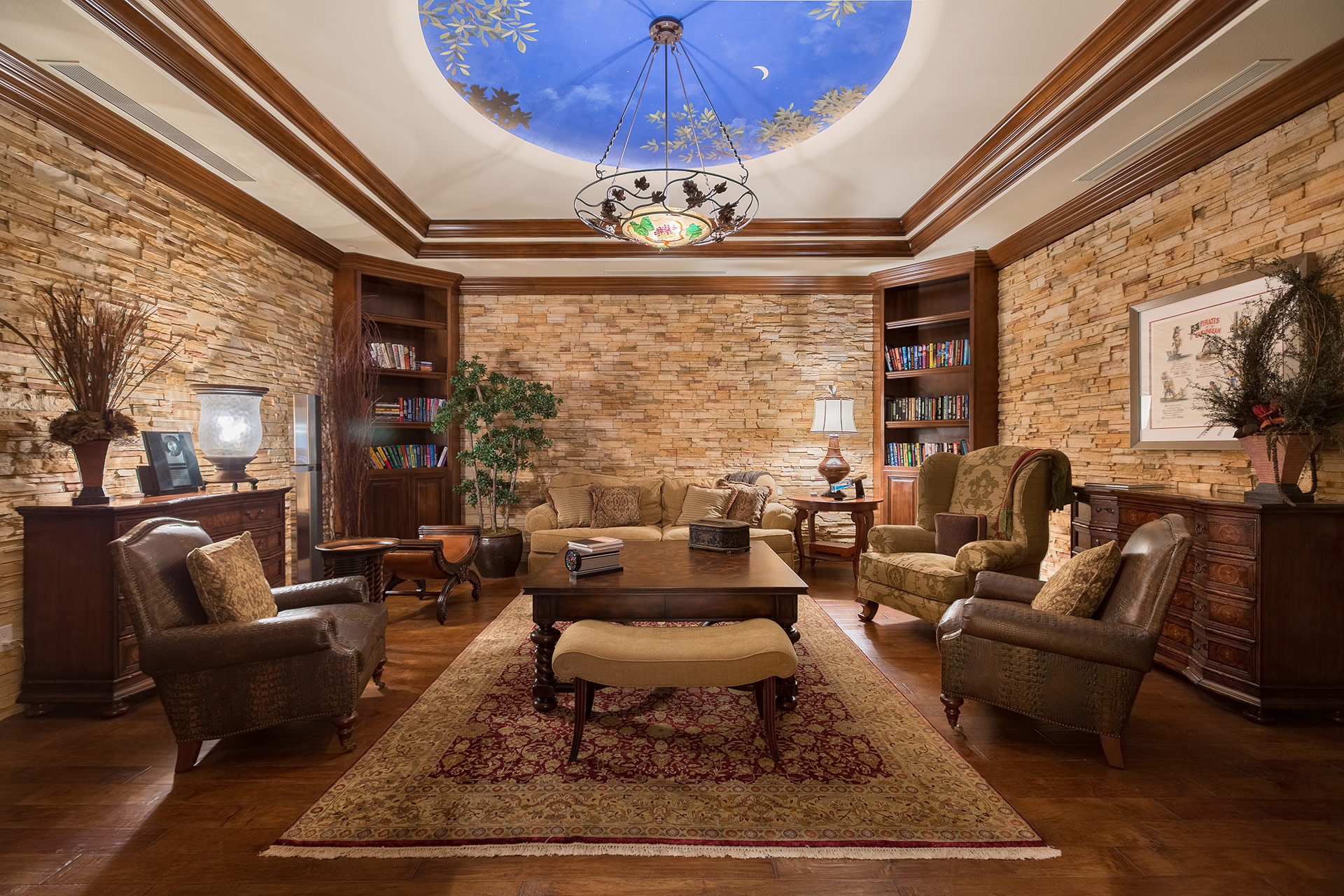 A dark, moody study with a recessed, blue and gold painted recessed medallion and chandelier on the ceiling, slate stone walls, 3 chairs, and dark-wood furniture.