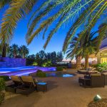 Palm fronds framing the top of a photo of a desert luxury home backyard at dusk with patio chairs, wading pool, lazy river and swimming pool, and view of Las Vegas in the background.