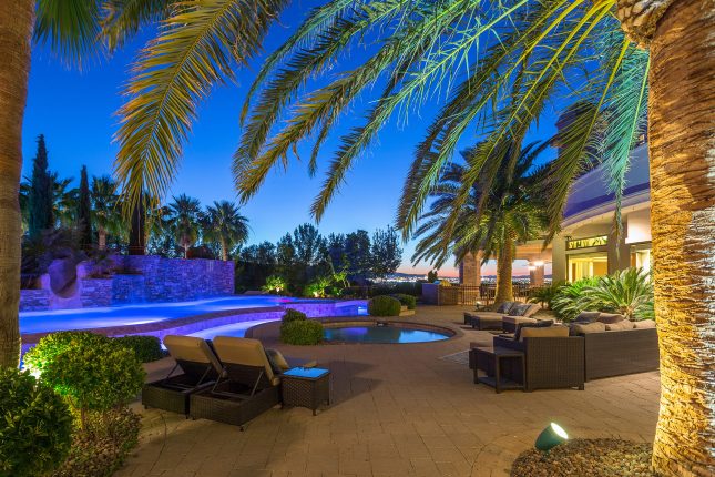 Palm fronds framing the top of a photo of a desert luxury home backyard at dusk with patio chairs, wading pool, lazy river and swimming pool, and view of Las Vegas in the background.