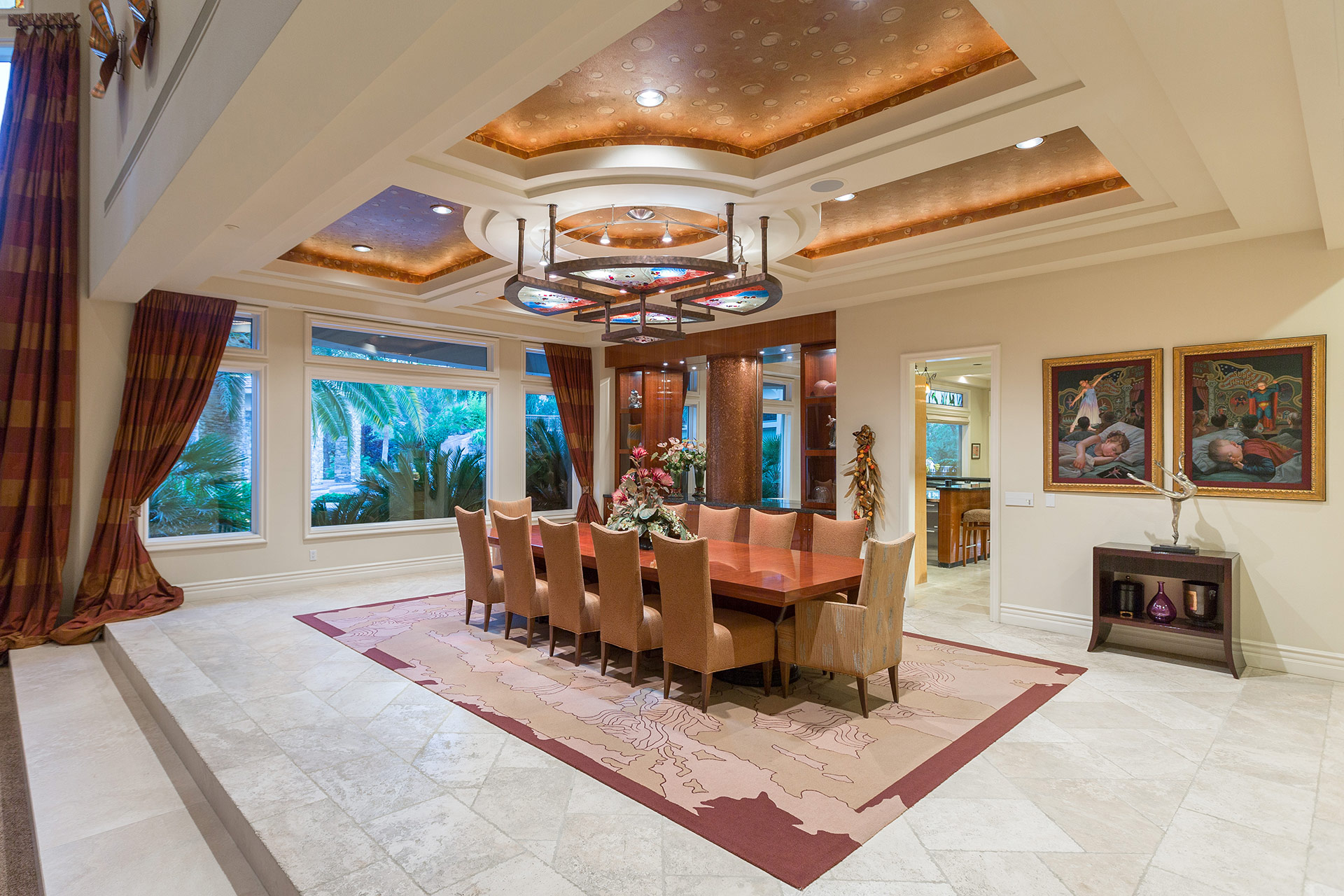 A large dining room with an area rug atop beige tiled floors, and large table with seating for twelve and a stained-glass light fixture hanging from a coffered ceiling with wall-papered inlay and recessed lighting.