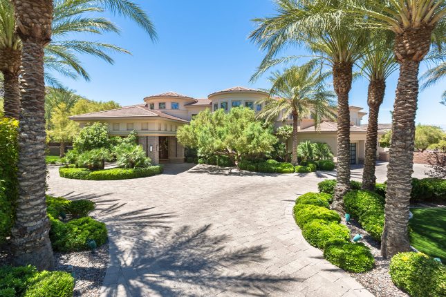 A driveway leading to a stucco-and-stone luxury home with porte-cochère, and two turrets, lined with palm trees and landscaped with trees and hedges.