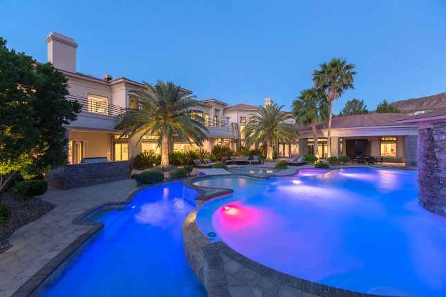 A luxury home backyard at twilight with swimming pool and lazy river in the foreground and palm trees, patio furniture and large home in the background.