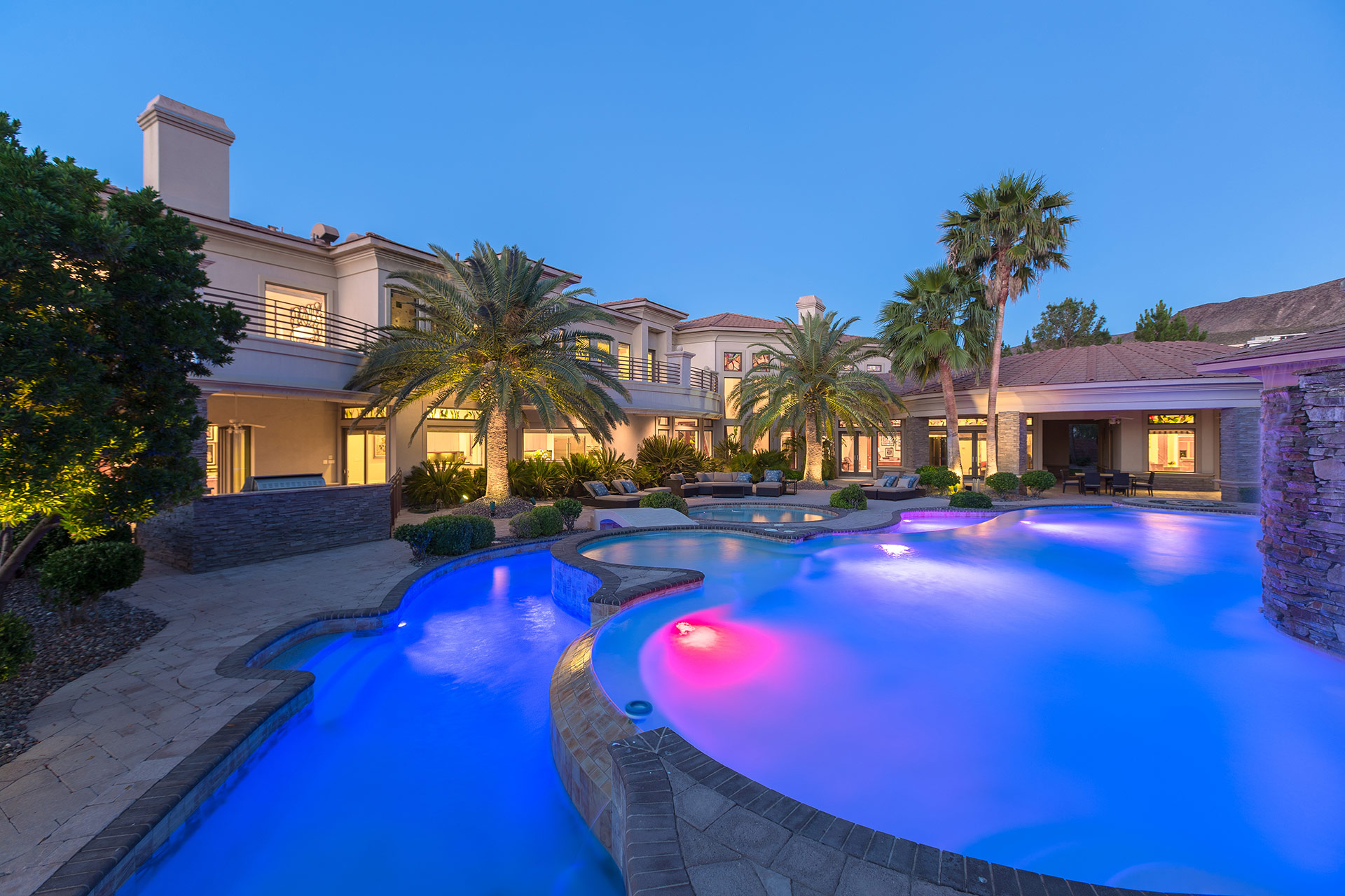 A luxury home backyard at twilight with swimming pool and lazy river in the foreground and palm trees, patio furniture and large home in the background.