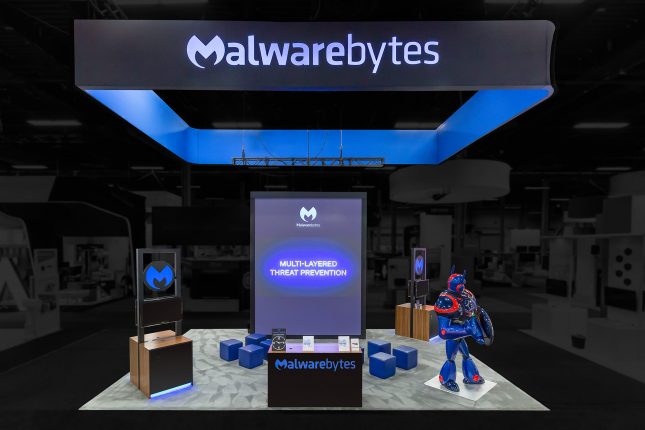 Malwarebytes trade show exhibit with a suspended black banner with rear-illuminated blue logo, and blue interior, a 15-foot tall video display, a black, wood-paneled welcome desk with blue logo on the front and two wood-finished kiosks within the exhibit and a 8-foot blue, red and black robot wielding a shield.