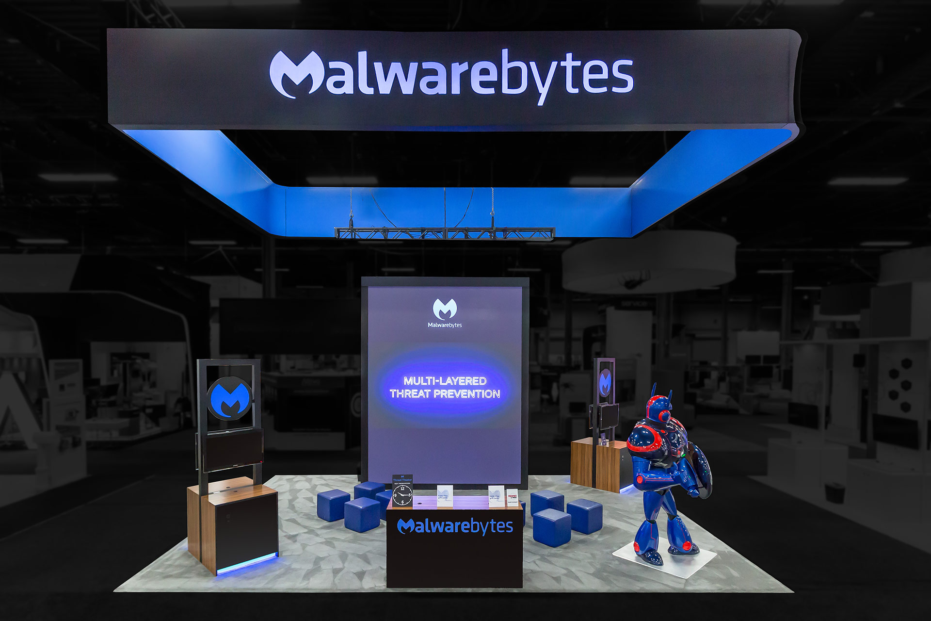 Malwarebytes trade show exhibit with a suspended black banner with rear-illuminated blue logo, and blue interior, a 15-foot tall video display, a black, wood-paneled welcome desk with blue logo on the front and two wood-finished kiosks within the exhibit and a 8-foot blue, red and black robot wielding a shield.
