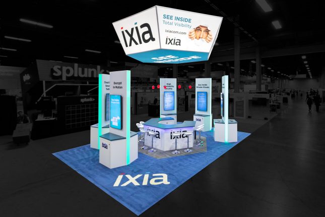 Elevated perspective of Ixia tradeshow exhibit with a white and blue suspended hexagonal banner above five white, tall stations with vertical video screens, a hexagonal counter in the center with modern white barstools and blue carpet with an Ixia logo.