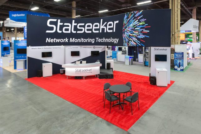 An elevated view of Statseeker trade show exhibit with a two-story display wall with Statseeker logo on dark blue background and white text above and video kiosks below. The exhibit has red carpet a grey desk in the foreground and a curved white counter in front of the wall.