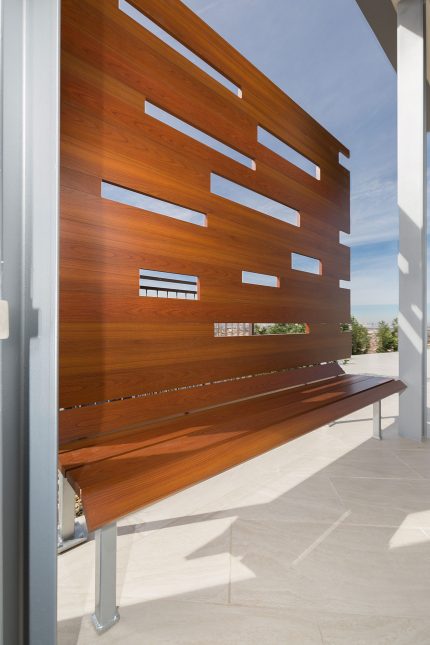 A wood-like aluminum backyard bench with its shadow on a light beige tiled patio with cloudy blue skies in the background.