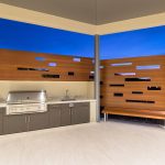 A stainless steel built-in backward bbq with wood-like aluminum fencing and bench atop light beige tiled flooring and a rich blue twilight sky in the background.