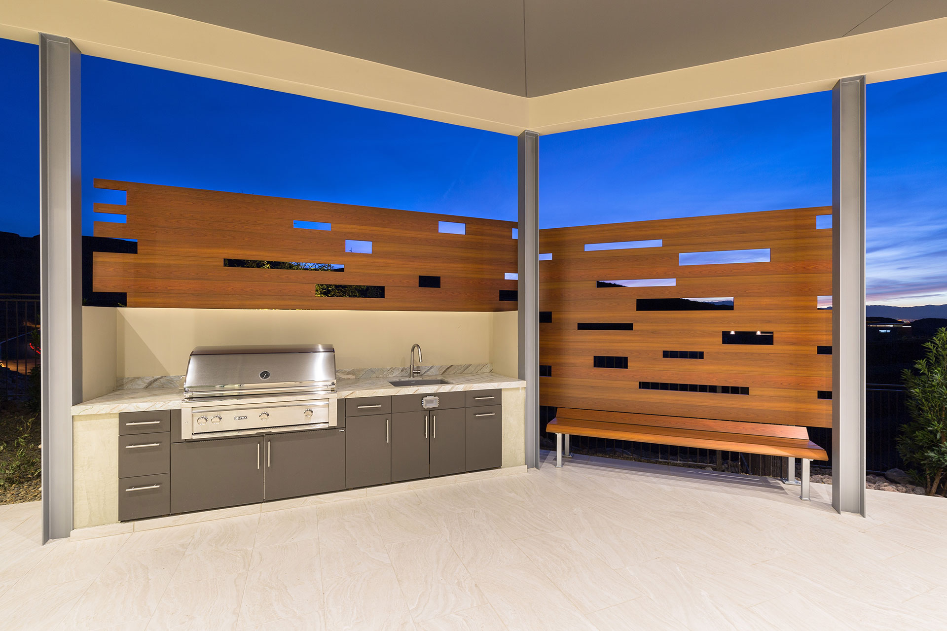 A stainless steel built-in backward bbq with wood-like aluminum fencing and bench atop light beige tiled flooring and a rich blue twilight sky in the background.