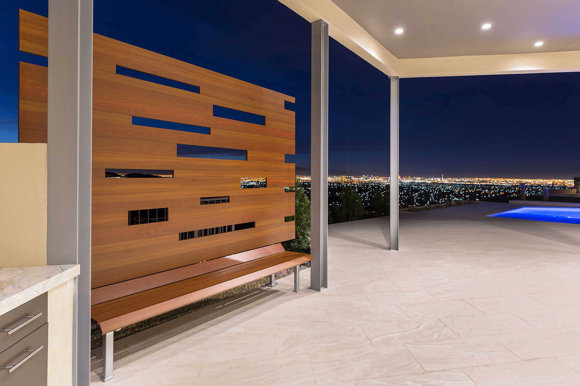 A wood-like aluminum backyard bench on a light beige tiled patio with Las Vegas city lights and a pool in the background.