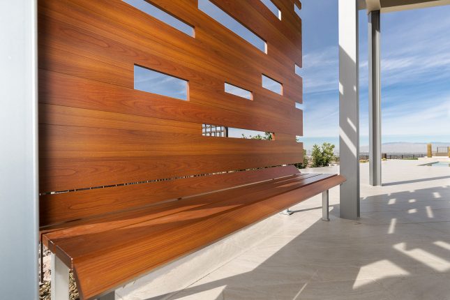 A wood-like aluminum backyard bench with its shadow on a light beige tiled patio with cloudy blue skies in the background.