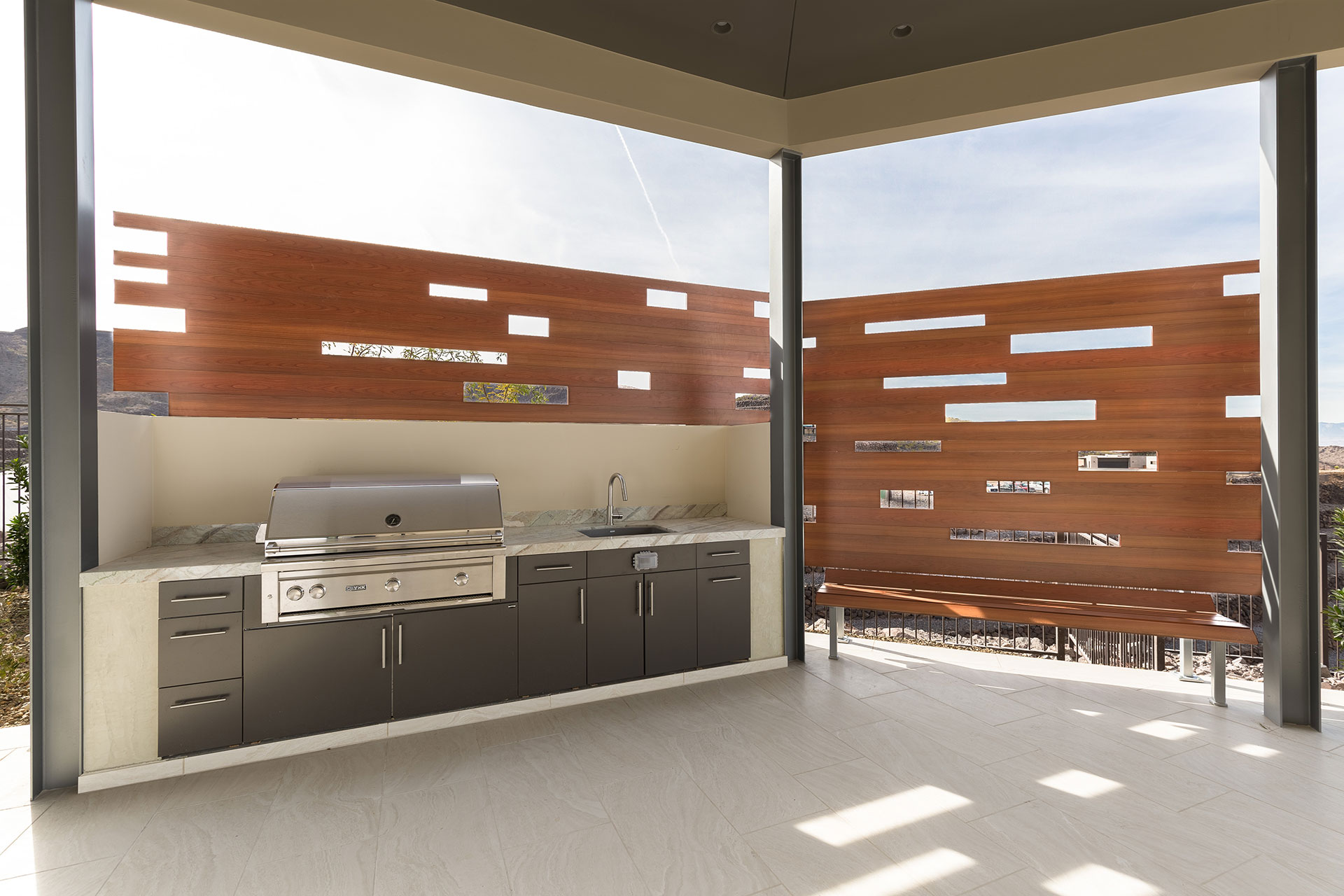 A stainless steel built-in backward bbq with wood-like aluminum fencing and bench atop light beige tiled flooring with cloudy bright daylight in the background.