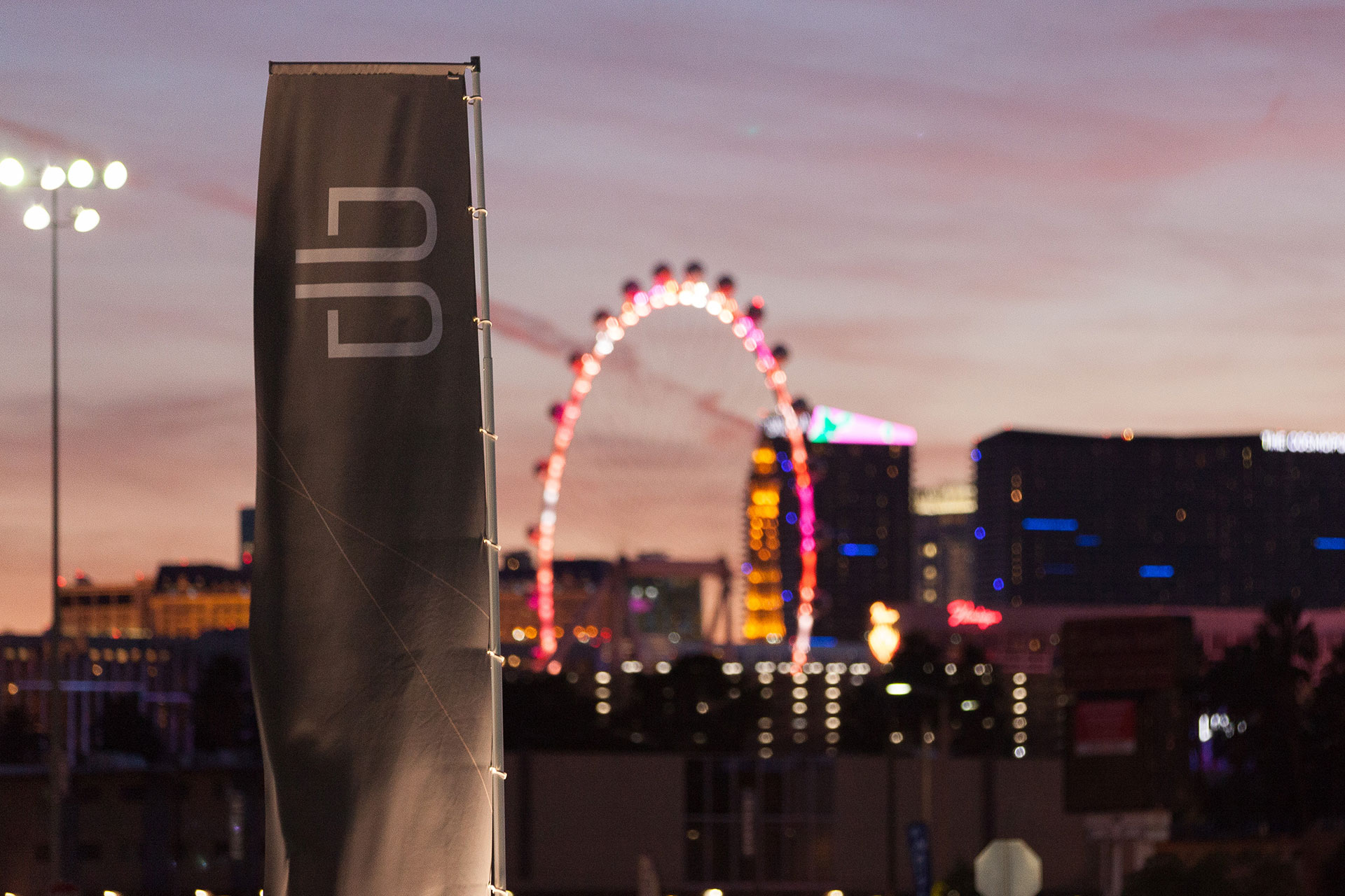 The white B of the Byton logo on a grey banner flapping in the wind in front of the High Roller Ferris Wheel and Casinos of the Las Vegas Strip at dusk.
