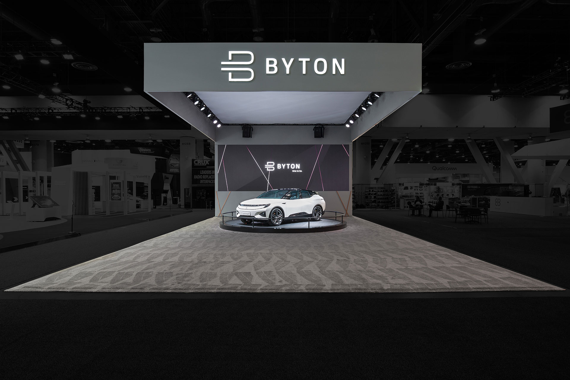 A wide-angle perspective of the Byton Electric Car CES Tradeshow Exhibit with a white automobile on a turntable on grey carpet with a white ceiling and grey walls and a white Byton logo above.