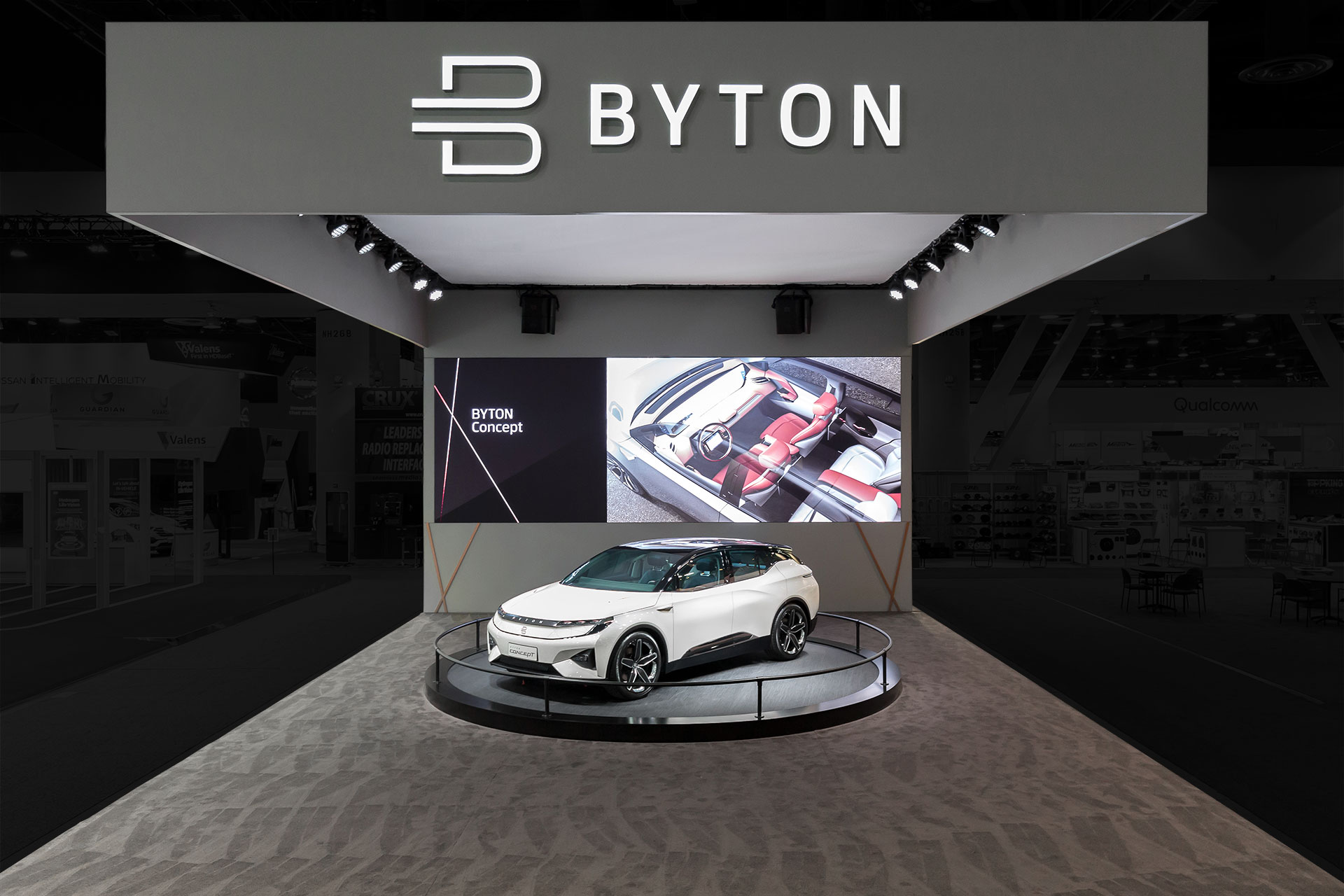 Byton Electric Car CES Tradeshow Exhibit with a white automobile on a turntable on grey carpet with a white ceiling and grey walls and a white Byton logo above.
