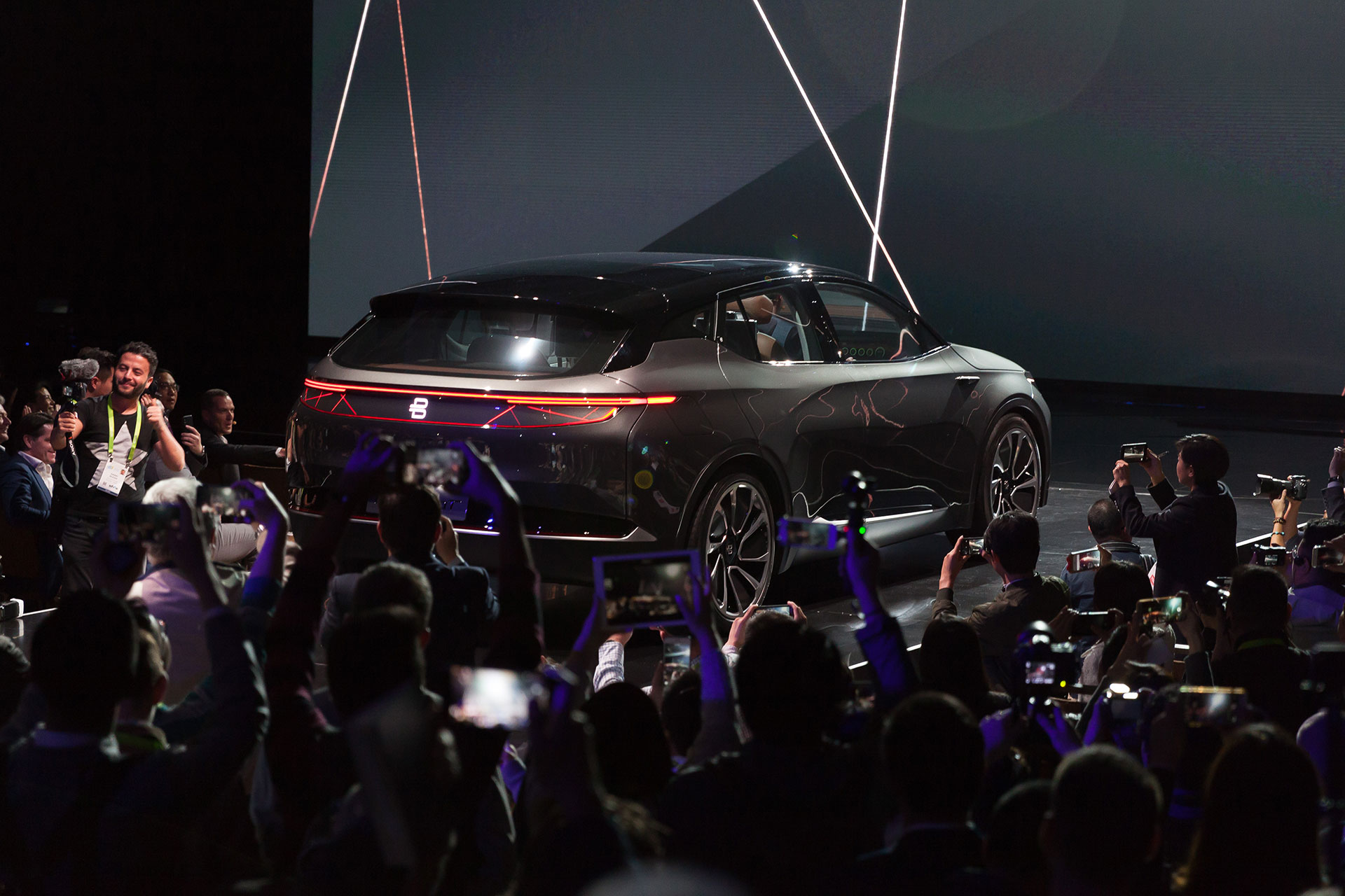A dark grey Byton concept car rolling down the runway at a press release swarmed by photographers.