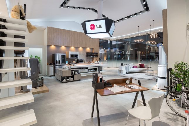 A light-colored home exhibit by LG with stairs on the left a small wooden desk in the foreground and a kitchen and sofa in the background.